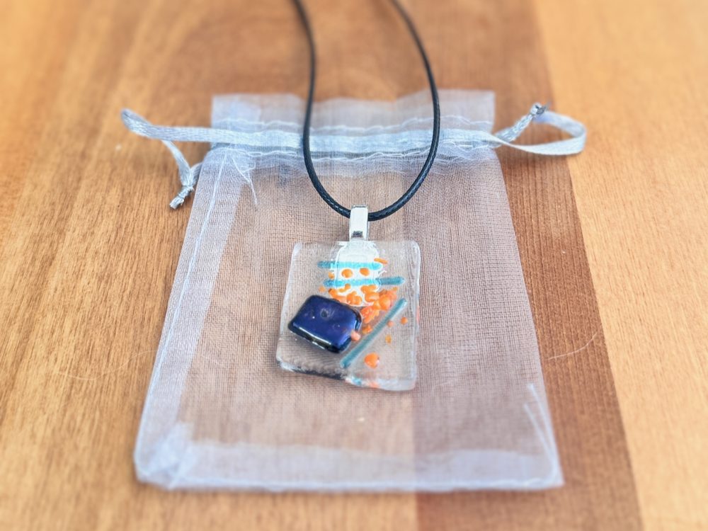 Tidal fused glass pendant in clear glass with a cobalt block, orange dots and turquoise lines, on black cord